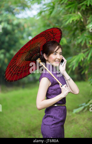 Chinesische Frau sprechen über ein Smart Phone und das Tragen eines Cheongsam chinesische Kleid mit traditionellen Regenschirm. Stockfoto