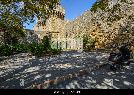 Eine schwarze und weiße stray Griechische Katze entspannt im Schatten der alten Burg von Rhodos Griechenland im Hintergrund und ein Motorroller Vespa in der Nähe Stockfoto