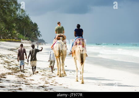 Zwei Touristen reiten auf Kamelen am Strand mit Einheimischen zu Fuß entlang der Seite, Diani, Kenia Stockfoto