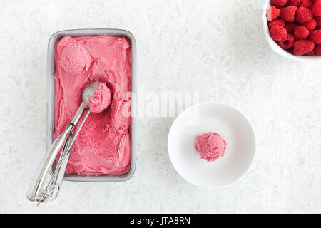 Schaufeln des hausgemachten Himbeer Eis in einem metallischen Whirlpool und weiße Schüssel zusammen mit frischen Himbeeren auf Weiß marmorierten Hintergrund angeordnet. Stockfoto