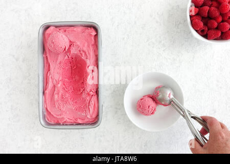 Hand Schaufeln aus einem Behälter mit hausgemachten Himbeer Eis gefüllt und eine Kugel in eine Schüssel. Stockfoto