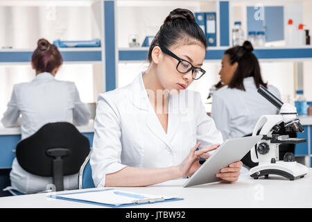 Asiatische Wissenschaftler in Laborkittel mit Mikroskop und digitale Tablette im chemischen Labor Wissenschaftler hinter Gruppe Stockfoto