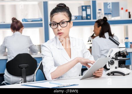 Asiatische Wissenschaftler in Laborkittel mit Mikroskop und digitale Tablette im chemischen Labor Wissenschaftler hinter Gruppe Stockfoto