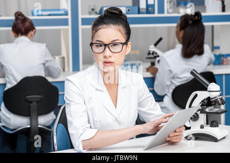 Asiatische Wissenschaftler in Laborkittel mit Mikroskop und digitale Tablette im chemischen Labor Wissenschaftler hinter Gruppe Stockfoto