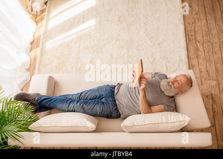 Älterer bärtiger Mann lächelnd und mit digitalen Tablet und liegend auf dem Sofa zu Hause Stockfoto