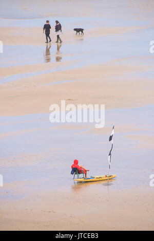 An einem ruhigen Strand einer einsamen RNLI Rettungsschwimmer in seinem Stuhl sitzt auf Pflicht als ein Paar vorbei mit Ihrem Hund. Stockfoto