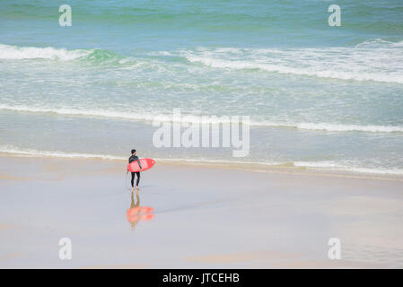 Ein einsamer Surfer mit einem roten Surfboard zu Fuß ins Meer. Newquay, Cornwall. Stockfoto