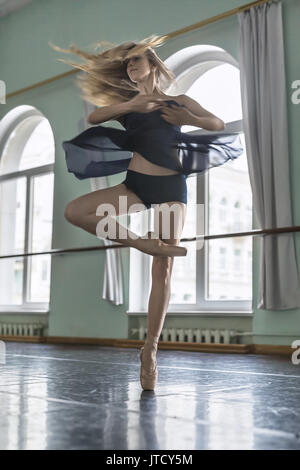 Junge Ballerina posiert in einer Motion im Ballett Halle gegenüber großen Bogen Windows. Sie kreisen auf dem linken Bein. Mädchen trägt einen dunklen Tanz tragen Sie ein Stockfoto