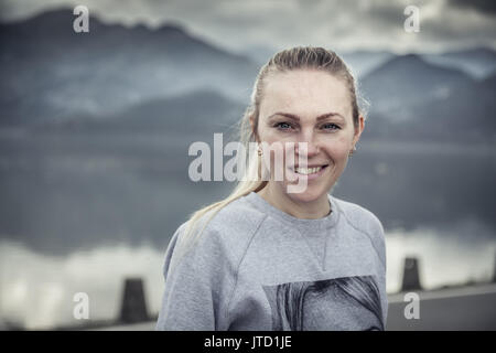 Blonde junge lächelnde Frau Porträt mit mit unscharfen Berge Landschaft auf dem Hintergrund im bewölkten Tag Stockfoto