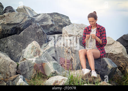 Eine schöne dunkelhaarige Frau in einem Plaid Shirt, denim Shorts, weiße Turnschuhe strickt mit Stricknadeln eine graue Strickjacke aus natürlichen woolen Gewinde Stockfoto