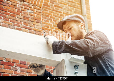 Ein starker Mann Builder in dunklen Bau Overalls und eine graue Kappe baut ein Haus von einer hölzernen Balken weiß gestrichen, an einem Sommertag, im Hintergrund ein Stockfoto