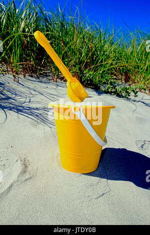 Ein Kind im gelben Eimer mit Sand und mit einer Schaufel an einem Strand gefüllt Stockfoto