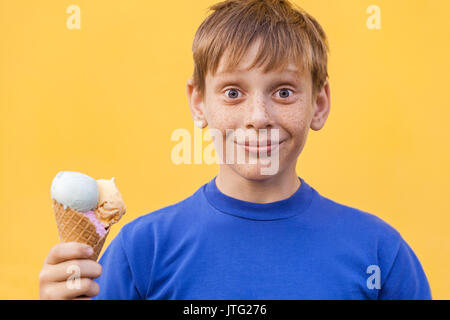 Blonde schöne Junge mit den Sommersprossen und blaues T-Shirt hält Eis am Kamera mit Lächeln und frage mich, Gesicht. Studio erschossen, auf einem gelben b isoliert Stockfoto