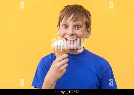 Die blonde schöne Junge mit den Sommersprossen und blaues T-Shirt hält Eis an Kamera suchen mit toothy Lächeln. Studio erschossen, auf einem gelben backgro isoliert Stockfoto