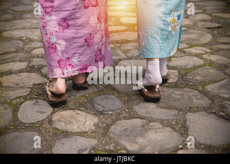 Closeup Foto der Rückseite traveler Freundinnen in traditioneller kimono Kleidung und japanische komaheta auf Stein Weg gemeinsam gehen mit Sonnenlicht Stockfoto