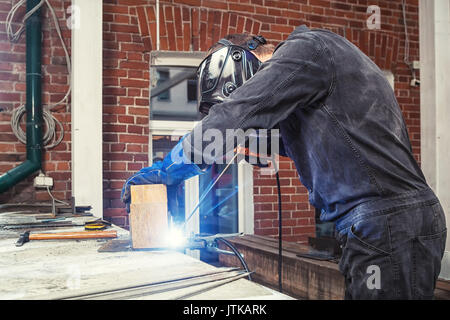 Eine starke männliche Schweißer auf einer Konstruktion schwarz insgesamt und schweißen Tupfer Schweißnähte ein Schweißgerät mit einer metallischen Struktur zu einer hölzernen Bar auf der Straße, in den Stockfoto