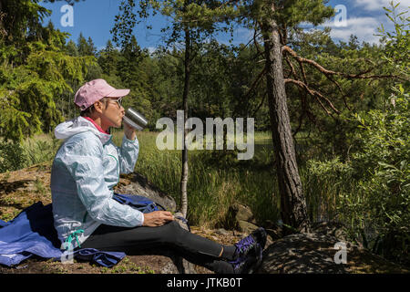 Reifen asiatischen Taiwanese chinesische Frau trinkt Kaffee in einer Pause während einer Wanderung um einen Wald See in Nynashamn, Schweden, an einem hellen Sommer Stockfoto
