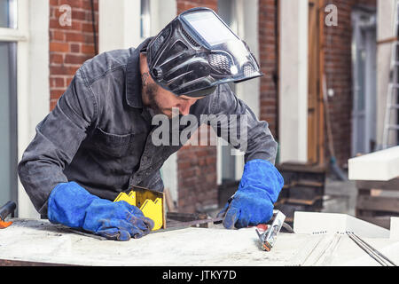 Ein Mann Builder in einem schweissverfahren Maske und blau Schutzhandschuhe macht eine Metallkonstruktion auf einem Schreibtisch auf einer Baustelle an einem Sommertag in den hinterg Stockfoto