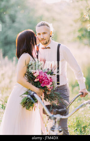 Die Nahaufnahme Porträt der Braut im weißen Brautkleid, die rosa Blumenstrauß und küssen die modern gekleidete Bräutigam mit dem Fahrrad in die Wange. Zusammensetzung im sonnigen Bereich. Stockfoto