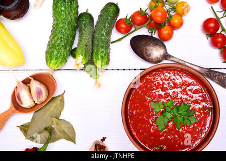 Frische gazpacho Suppe in eine braune Keramik Teller mit einem Löffel auf einer weißen Holz- Hintergrund, neben frischem Gemüse und Gewürze zum Kochen, Ansicht von oben Stockfoto