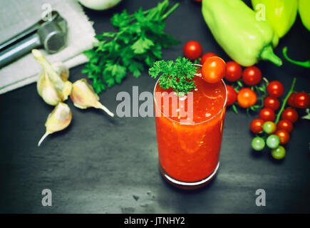 Frisch zubereiteten Saft aus Tomaten und Gewürzen in ein Glas auf einem schwarzen Hintergrund, Ansicht von oben, hinter frischem Gemüse Stockfoto