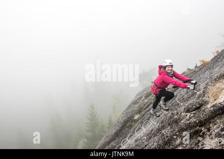 Eine Frau lächelt mit Aufregung, wie sie klettert das Metall Sprossen einer Klettersteig in Squamish. Stockfoto