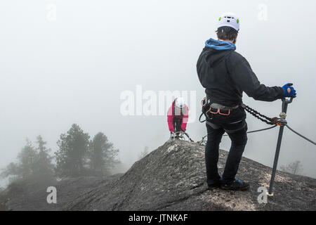 Ein Mann wartet und schaut zurück als Frauen klettert die Metall Sprossen einer Klettersteig in Squamish, British Columbia. Stockfoto