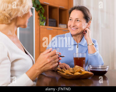 Zwei lächelnde positive weibliche Rentner trinken Kaffee am Tisch zu Hause Stockfoto