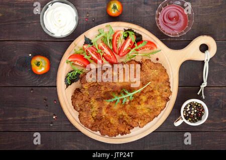 Eine große Wiener Schnitzel und Salat auf einem Schneidebrett auf einem dunklen Hintergrund. Ansicht von oben. Stockfoto