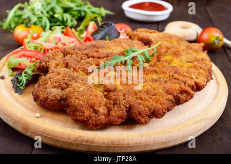 Eine große Wiener Schnitzel und Salat auf einem Schneidebrett auf einem dunklen Hintergrund. Nahaufnahme Stockfoto