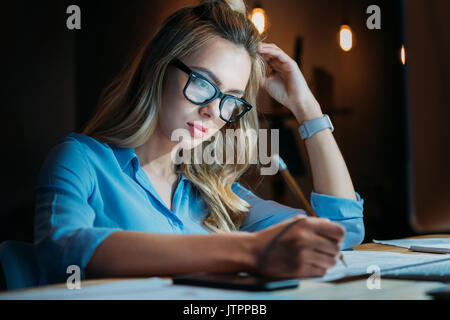 Müde blond kaukasischen Student in Brillen etwas Schreiben mit Bleistift und Studieren bis spät Stockfoto