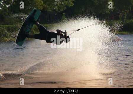 Wake Boarder in Mid-Air tun Half-Somersault auf See in der Nähe der Sonnenuntergang hinter Plume von Spritzwasser Stockfoto