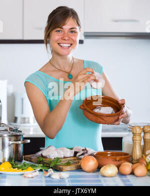 Fröhlicher junger Hausfrau kochen Seehecht in Keramik Topf zu Hause Stockfoto