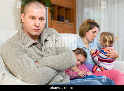 Frau kann nicht seinen Mann vergeben. Man sitzt auf einem Sofa auf der Vorderseite und seiner Familie sitzt hinter ihm. Zwei weinenden Töchter und einen enttäuscht wi Stockfoto