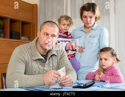 Familie Probleme mit Bargeld. Vater sitzt am Tisch mit Geld in den Händen seiner Frau und seinen zwei Töchtern traurig hinter ihm sitzen Stockfoto