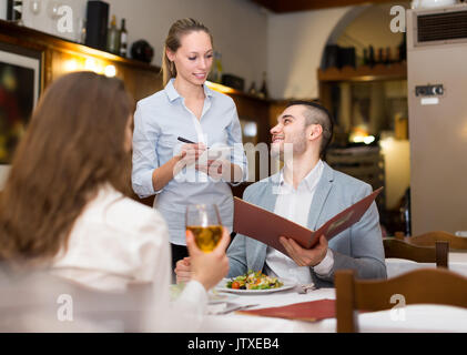 Junge blonde Kellnerin mit leckeren Gerichten an zufriedene Gäste Paar. Selektiver Fokus Stockfoto