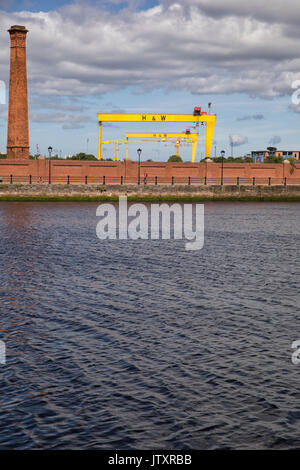 Belfast, ein Blick auf die alte Werft Harland und Wolff Krane (Samson und Goliath) mit Fluss Lagan, Schornstein und Red brick wall im Vordergrund Stockfoto
