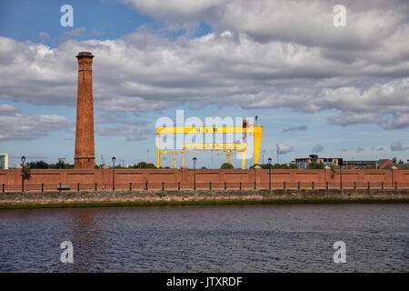 Belfast, ein Blick auf die alte Werft Harland und Wolff Krane (Samson und Goliath) mit Fluss Lagan, Schornstein und Red brick wall im Vordergrund Stockfoto