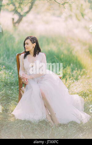 Porträt der schönen Frau in der langen weißen Kleid und mit dem Zubehör auf Ihrem Haar woanders suchen und Sitzen im Alten moder Stuhl in das Feld ein. Stockfoto