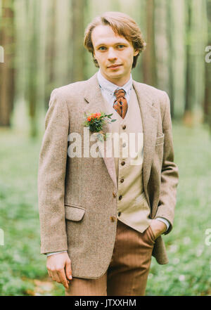 Mann in der vintage Anzug, mit Mini-Blumenstrauß ist auf der Jacke befestigt ist mit Blick auf die Kamera und halten die Hand in der Tasche. Die close-up front Porträt in den Wald. Stockfoto