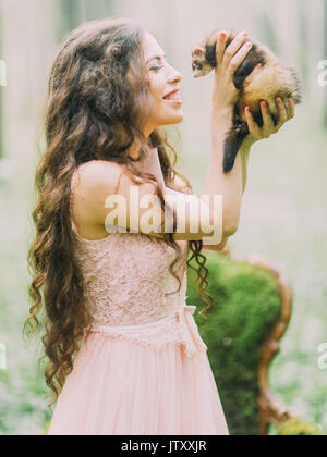 Die glückliche Frau mit langen lockigen Haar im Lichte rosa Kleid Holding, Schmusen und Spielen mit den Frettchen in den grünen Wald. Stockfoto