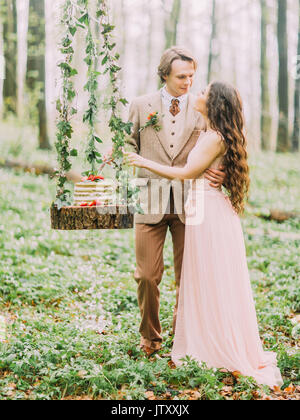 Die vertikale Foto der Jungvermählten stehen in der Nähe des hängenden Stumpf mit der Hochzeit in Grün und Weiß Kuchen auf Sie. Die glücklich gerade im grünen Frühling Wald verheiratet. Stockfoto