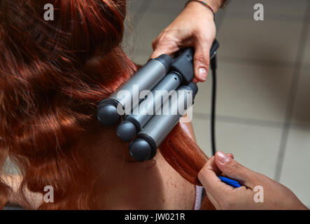 Junge rothaarige Frau, die ihr Haar gewelltes mit modernen elektrischen Triple barrel Lockenstab in ein Haar styling Salon. Detailansicht. Stockfoto
