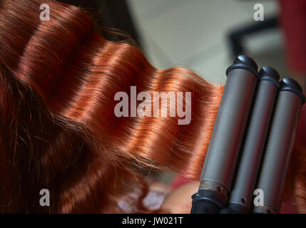 Nahaufnahme der Lange rote Haare kräuselten sich mit elektrischen Triple barrel Lockenstab in einen Friseursalon. Stockfoto