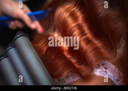 Junge Frau ihre weiche rötliche Haare in Geringelt mit elektrischen Triple barrel Lockenstab in einen Friseursalon. closeup. Stockfoto