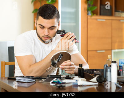 Überzeugter Kerl rasieren elektrische Rasierer zu Hause sitzen an einem Tisch im Wohnzimmer Stockfoto