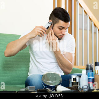 Schöner Mann rasieren elektrische Rasierer zu Hause sitzen auf einem Sofa im Wohnzimmer Stockfoto