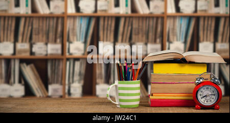 Stapel von Büchern Becher mit Buntstiften und Wecker auf hölzernen Tisch gegen verschiedene Mehrfarbige Bücher im Regal Stockfoto