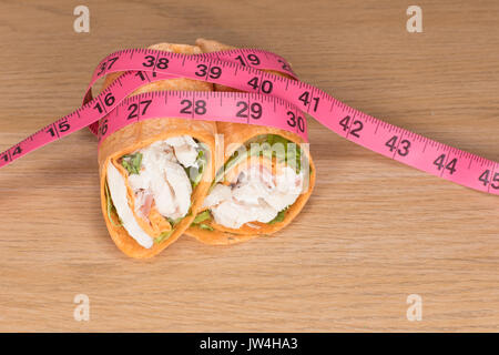 Ein Huhn, Salat wrap geschnitten in der Hälfte mit einem rosa Maßband auf einer Ebene Holz Tisch Hintergrund in einem Gewichtsverlust und Diät Konzept. Stockfoto