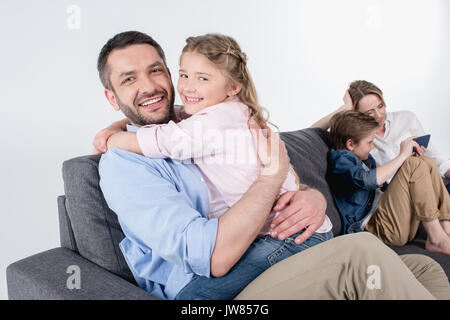 Vater mit Tochter umarmen und auf Kamera, während Mutter mit Sohn sitzen im Sofa Stockfoto